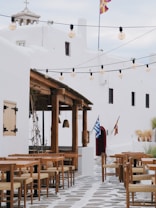 A picturesque outdoor cafe setting with wooden tables and chairs arranged neatly on a stone-paved walkway. Above, string lights hang across the scene, adding charm. In the background, there is a whitewashed building with traditional architecture, featuring small windows and a rooftop cross, indicative of a church. Two flags are visible, one with blue and white stripes and another with a different design. The area is adorned with potted plants, adding greenery to the serene setting.