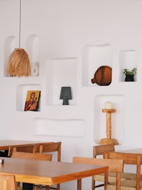 A minimalist interior is characterized by white walls with recessed shelves displaying various decorative items. The shelves hold objects such as a woven lampshade, a religious icon painting, a wooden board, a potted plant in a black vase, and a carved candle holder with a white candle. The space is furnished with simple wooden tables and chairs, creating an inviting atmosphere.