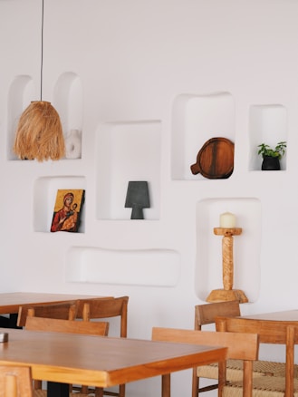 A minimalist interior is characterized by white walls with recessed shelves displaying various decorative items. The shelves hold objects such as a woven lampshade, a religious icon painting, a wooden board, a potted plant in a black vase, and a carved candle holder with a white candle. The space is furnished with simple wooden tables and chairs, creating an inviting atmosphere.