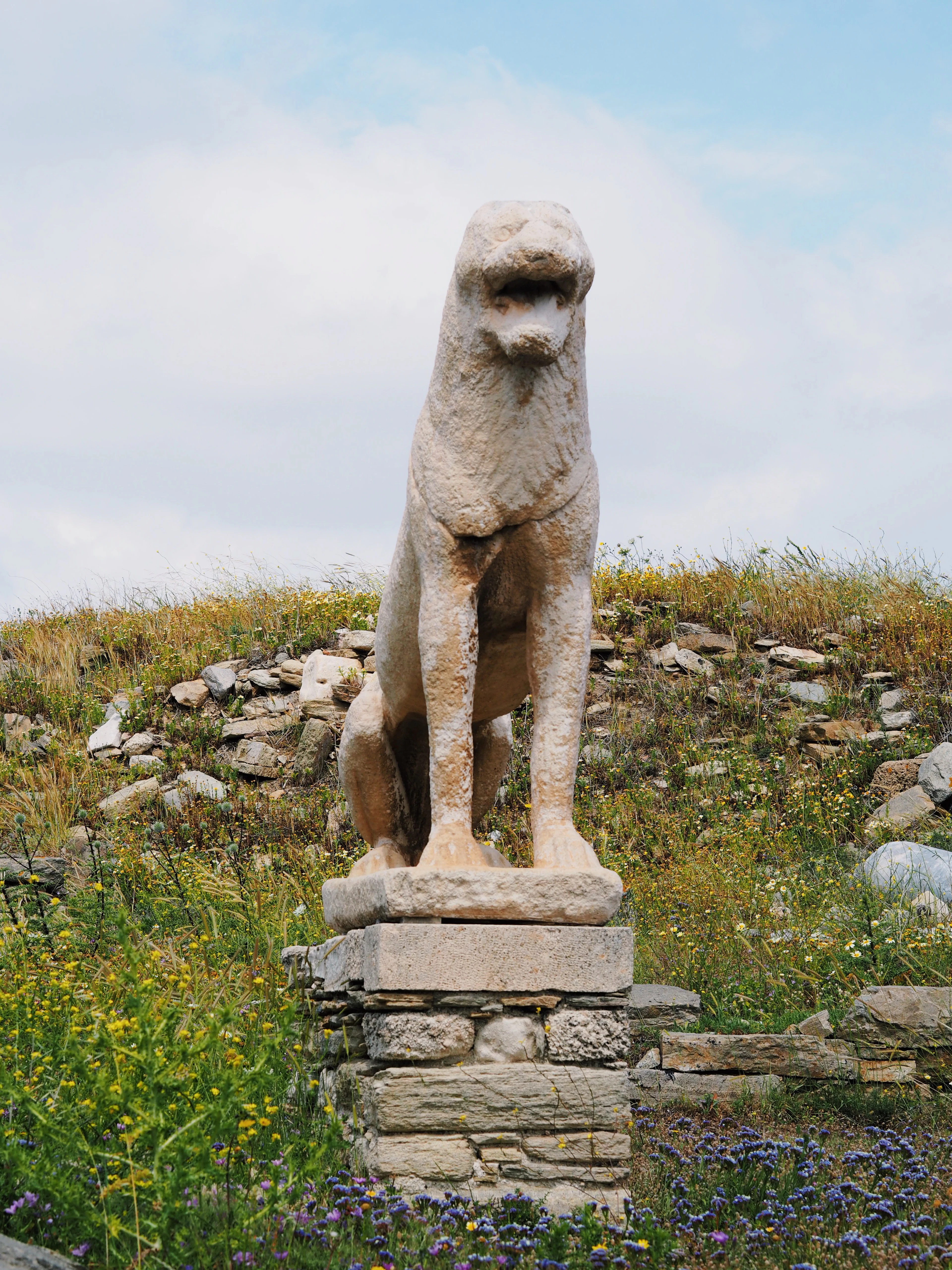 One of the archaic marble lions of Delos sitting upright on its plinth above the excavated stones of the sanctuary, surrounded by yellow and violet spring wildflowers.