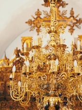 A highly ornate and intricate chandelier made of polished gold-colored metal with elaborate scrollwork and multiple candle-shaped light bulbs. The background features a luxurious interior with detailed gold decorations and motifs.