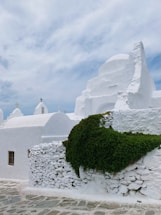 A whitewashed building with rounded architectural features and small, simple crosses on top, typical of Cycladic architecture. A dense patch of vibrant green ivy covers part of the stone wall. The sky is partly cloudy, creating a serene and peaceful atmosphere.