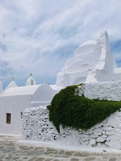 A whitewashed building with rounded architectural features and small, simple crosses on top, typical of Cycladic architecture. A dense patch of vibrant green ivy covers part of the stone wall. The sky is partly cloudy, creating a serene and peaceful atmosphere.