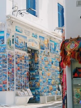 A vibrant street-side shop displays an extensive collection of colorful postcards featuring scenic images of Mykonos and other Greek landmarks. The storefront is adorned with blue accents, matching the blue shutters of the building. To the right, a variety of woven bags and textiles hang, adding a splash of red and orange to the scene.