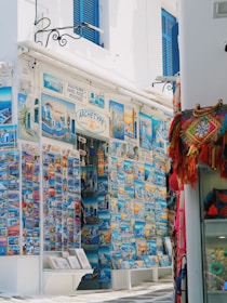 A vibrant street-side shop displays an extensive collection of colorful postcards featuring scenic images of Mykonos and other Greek landmarks. The storefront is adorned with blue accents, matching the blue shutters of the building. To the right, a variety of woven bags and textiles hang, adding a splash of red and orange to the scene.