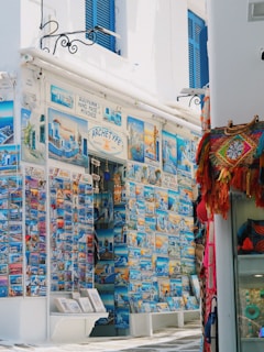 A vibrant street-side shop displays an extensive collection of colorful postcards featuring scenic images of Mykonos and other Greek landmarks. The storefront is adorned with blue accents, matching the blue shutters of the building. To the right, a variety of woven bags and textiles hang, adding a splash of red and orange to the scene.