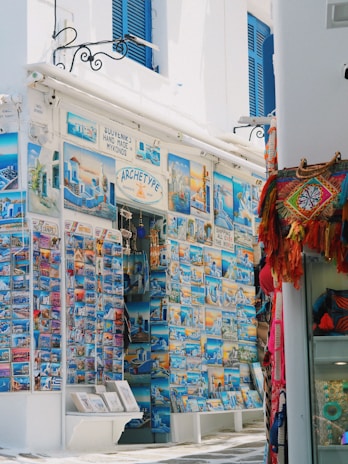 A vibrant street-side shop displays an extensive collection of colorful postcards featuring scenic images of Mykonos and other Greek landmarks. The storefront is adorned with blue accents, matching the blue shutters of the building. To the right, a variety of woven bags and textiles hang, adding a splash of red and orange to the scene.