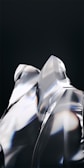 Smooth, reflective surfaces with sharp angles and edges create a glass-like appearance. Light refracts through the transparent material, casting subtle rainbow-like hues against a dark background. The composition appears abstract and modern, emphasizing form and light interaction.