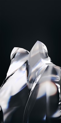 Smooth, reflective surfaces with sharp angles and edges create a glass-like appearance. Light refracts through the transparent material, casting subtle rainbow-like hues against a dark background. The composition appears abstract and modern, emphasizing form and light interaction.