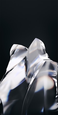 Smooth, reflective surfaces with sharp angles and edges create a glass-like appearance. Light refracts through the transparent material, casting subtle rainbow-like hues against a dark background. The composition appears abstract and modern, emphasizing form and light interaction.