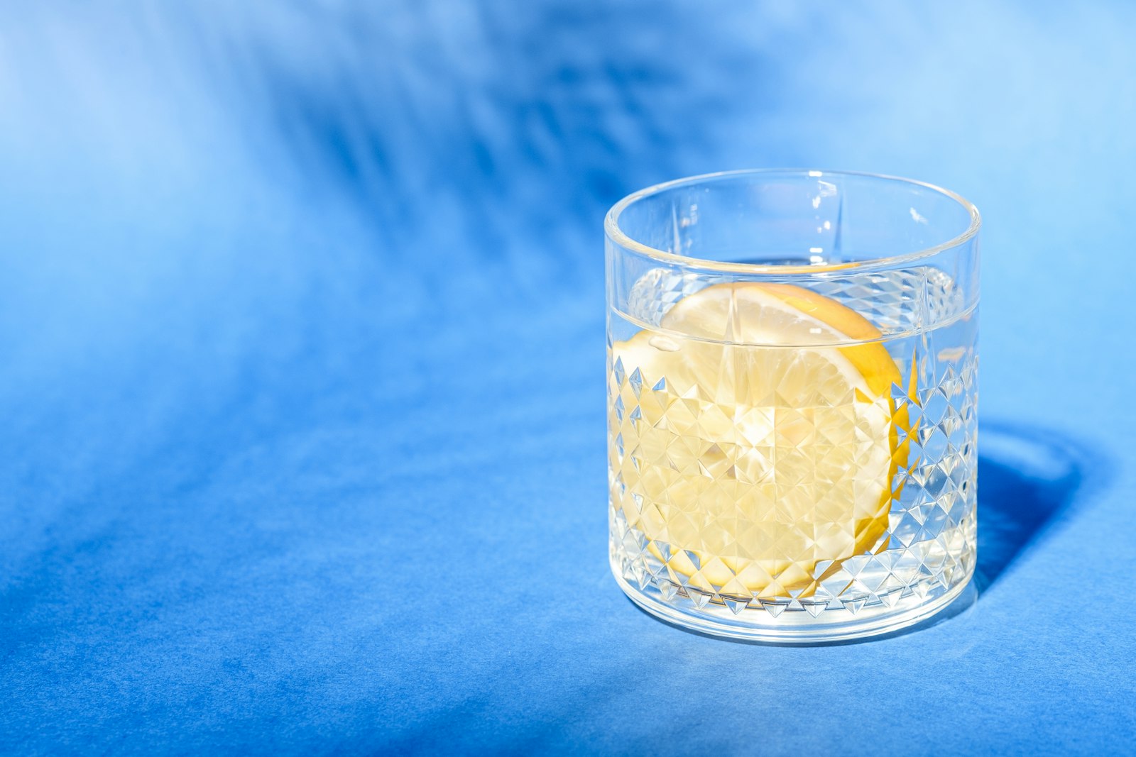 A glass of water with a slice of lemon on a blue surface in sunlight