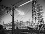 A complex structure of electricity pylons and wires extends across the landscape, with several large towers linked by numerous cables. The scene appears industrial and is dominated by the intricate metal frameworks of the towers and the crisscrossing wires.
