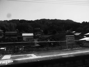 A railway platform surrounded by rural buildings and a lush, tree-covered hill in the background. There's a sign on the platform with Japanese writing.