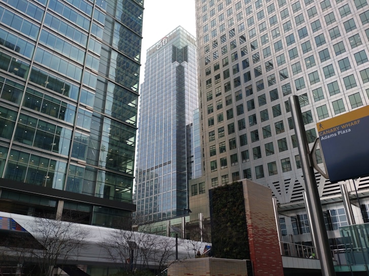 A cityscape featuring tall modern office buildings with large glass windows and reflective surfaces. A prominent sign near the bottom right corner reads 'Canary Wharf Adams Plaza'. Trees without leaves are visible in the foreground, along with a wall covered in greenery adding a touch of nature to the urban environment.