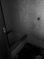 A small, dimly lit room with tiled walls surrounding a rectangular recess in the floor, possibly an old or neglected bathtub. The floor appears cluttered with debris and scattered objects, giving an impression of abandonment.
