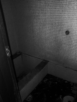 A small, dimly lit room with tiled walls surrounding a rectangular recess in the floor, possibly an old or neglected bathtub. The floor appears cluttered with debris and scattered objects, giving an impression of abandonment.