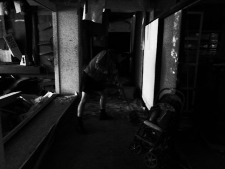 A person in a dimly lit, cluttered room seems to be engaged in cleaning or organizing. Debris and various items are scattered around, along with what appears to be a stroller on the right. The overall setting has a feeling of neglect or disarray.