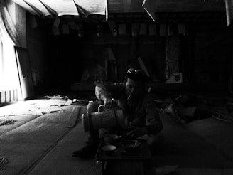 A person is seated in a dimly lit, cluttered room, pouring a liquid from a large container into a bowl while wearing sunglasses. The room is filled with scattered papers and decorative items hanging from the ceiling. Light enters through a window on the left, casting shadows in the space.