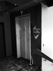 A dilapidated and abandoned interior of a building, featuring an elevator door on the seventh floor. Debris and peeling paint cover the floor, and there are visible signs of decay on the walls and ceiling.