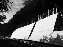 A concrete retaining wall runs alongside a road, bordered by a fence on top. The wall casts pronounced shadows due to the angle of sunlight, and the road curves into the distance. Trees with dense foliage surround the scene, creating a contrast with the bright surface of the wall.