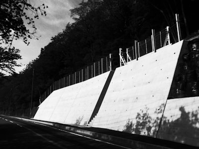 A concrete retaining wall runs alongside a road, bordered by a fence on top. The wall casts pronounced shadows due to the angle of sunlight, and the road curves into the distance. Trees with dense foliage surround the scene, creating a contrast with the bright surface of the wall.