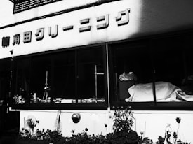A monochrome image of a building exterior with large windows. Japanese text is visible above the windows, likely indicating the name of a business or establishment. Inside the windows, various items and possibly furniture are visible, partially covered by a cloth. The lower part of the building has plants and flowers.