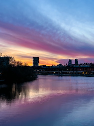 The image features a serene sunset over a cityscape, with silhouettes of buildings against a vibrant sky. The sky displays a gradient of colors from deep blue to shades of pink, purple, and orange, reflecting on a calm body of water in the foreground. Dark foliage lines the left edge, enhancing the contrast with the bright sky.