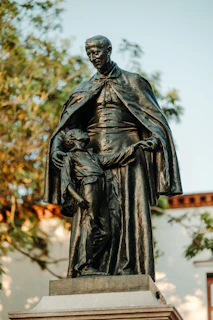 A bronze statue depicts a robed man standing with a smaller figure, possibly a child, who is grasping the man's cloak. They are depicted in a nurturing or protective pose, positioned on a pedestal amidst a backdrop of trees.