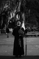 A person wearing a long black robe with a hood stands facing a rocky grotto. They have a backpack slung over one shoulder. In the background, a circular candleholder is lit, and the faint outline of a statue is visible within the grotto. The scene is captured in black and white, creating a solemn atmosphere.