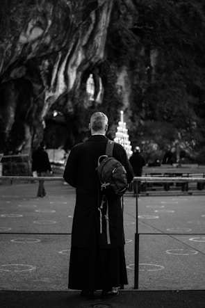A person wearing a long black robe with a hood stands facing a rocky grotto. They have a backpack slung over one shoulder. In the background, a circular candleholder is lit, and the faint outline of a statue is visible within the grotto. The scene is captured in black and white, creating a solemn atmosphere.