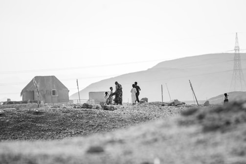 A black and white image showing a rural landscape with a simple dwelling made of mud and thatched roof on the left. Several people, including children and adults, are walking around the rocky ground. In the distance, there are power lines and a tall electricity pylon against a backdrop of hills.