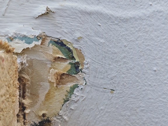 A close-up of a damaged wall surface showing a layer of peeling paint. The texture includes frayed and rough edges, with some areas revealing underlying wood or plaster. The surface appears worn and scratched.