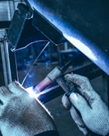 A person wearing knitted gloves is performing a welding task on a metal piece. Sparks and a bright arc light are visible at the welding point, with a backdrop of industrial equipment in the background.