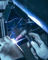 A person wearing knitted gloves is performing a welding task on a metal piece. Sparks and a bright arc light are visible at the welding point, with a backdrop of industrial equipment in the background.