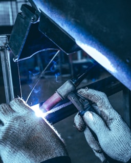 A person wearing knitted gloves is performing a welding task on a metal piece. Sparks and a bright arc light are visible at the welding point, with a backdrop of industrial equipment in the background.