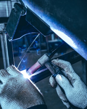 A person wearing knitted gloves is performing a welding task on a metal piece. Sparks and a bright arc light are visible at the welding point, with a backdrop of industrial equipment in the background.