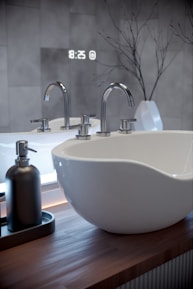 A modern bathroom sink with sleek chrome faucets atop a wooden countertop. A soap dispenser is placed to the left. In the background, a digital clock reads 18:25, and a white vase holds bare branch decor against a tiled wall.