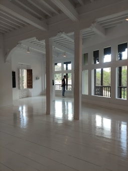 A spacious, bright room with large windows, white wooden floors, and white beams on the ceiling. Sunlight illuminates the space, which is minimalistic and modern. A person is standing by the windows, looking outside.