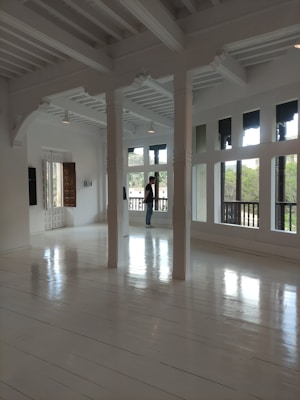 A spacious, bright room with large windows, white wooden floors, and white beams on the ceiling. Sunlight illuminates the space, which is minimalistic and modern. A person is standing by the windows, looking outside.