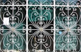 Ornate metal grilles with intricate swirling patterns cover three tall windows. Each section includes floral motifs at the center, framed by delicate, curving bars that create symmetrical designs. Behind the grilles, semi-transparent window blinds are visible, suggesting an interior setting beyond the decorative exterior.