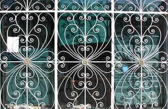 Ornate metal grilles with intricate swirling patterns cover three tall windows. Each section includes floral motifs at the center, framed by delicate, curving bars that create symmetrical designs. Behind the grilles, semi-transparent window blinds are visible, suggesting an interior setting beyond the decorative exterior.