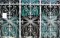 Ornate metal grilles with intricate swirling patterns cover three tall windows. Each section includes floral motifs at the center, framed by delicate, curving bars that create symmetrical designs. Behind the grilles, semi-transparent window blinds are visible, suggesting an interior setting beyond the decorative exterior.