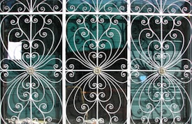 Ornate metal grilles with intricate swirling patterns cover three tall windows. Each section includes floral motifs at the center, framed by delicate, curving bars that create symmetrical designs. Behind the grilles, semi-transparent window blinds are visible, suggesting an interior setting beyond the decorative exterior.