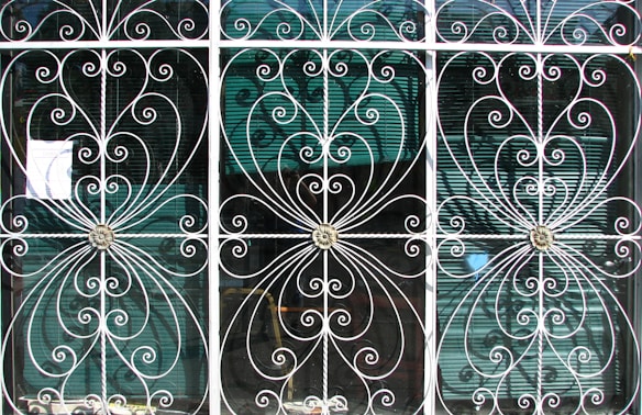 Ornate metal grilles with intricate swirling patterns cover three tall windows. Each section includes floral motifs at the center, framed by delicate, curving bars that create symmetrical designs. Behind the grilles, semi-transparent window blinds are visible, suggesting an interior setting beyond the decorative exterior.