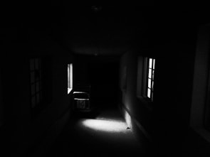 A dimly lit corridor with two windows on the right side, emitting faint light. A ghostly metal frame or cart sits against one of the windows, casting shadows on the floor. The atmosphere is dark and eerie.