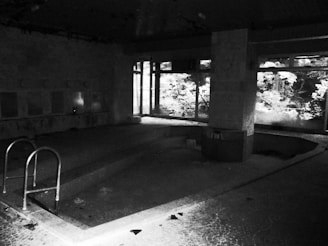 A dimly lit, abandoned indoor swimming pool with broken tiles and debris scattered around. The ceiling appears to have some decay, with peeling paint. Large windows on one side allow some light to filter in, casting shadows and partially illuminating the remaining features of the pool area.