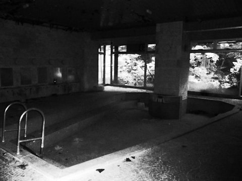 A dimly lit, abandoned indoor swimming pool with broken tiles and debris scattered around. The ceiling appears to have some decay, with peeling paint. Large windows on one side allow some light to filter in, casting shadows and partially illuminating the remaining features of the pool area.