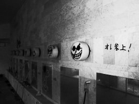 Graffiti with cartoon faces and bold Japanese characters adorns a wall in a dimly lit area. The faces have exaggerated features and are wearing sunglasses, creating a playful yet eerie vibe. The setting appears to be an industrial or public space with visible fixtures.