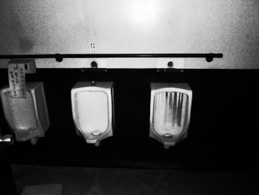 A black and white image featuring three urinals mounted on a tiled wall. The urinals are placed beneath a long horizontal bar with hooks. One urinal has a piece of paper with text attached to it.