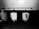 A black and white image featuring three urinals mounted on a tiled wall. The urinals are placed beneath a long horizontal bar with hooks. One urinal has a piece of paper with text attached to it.